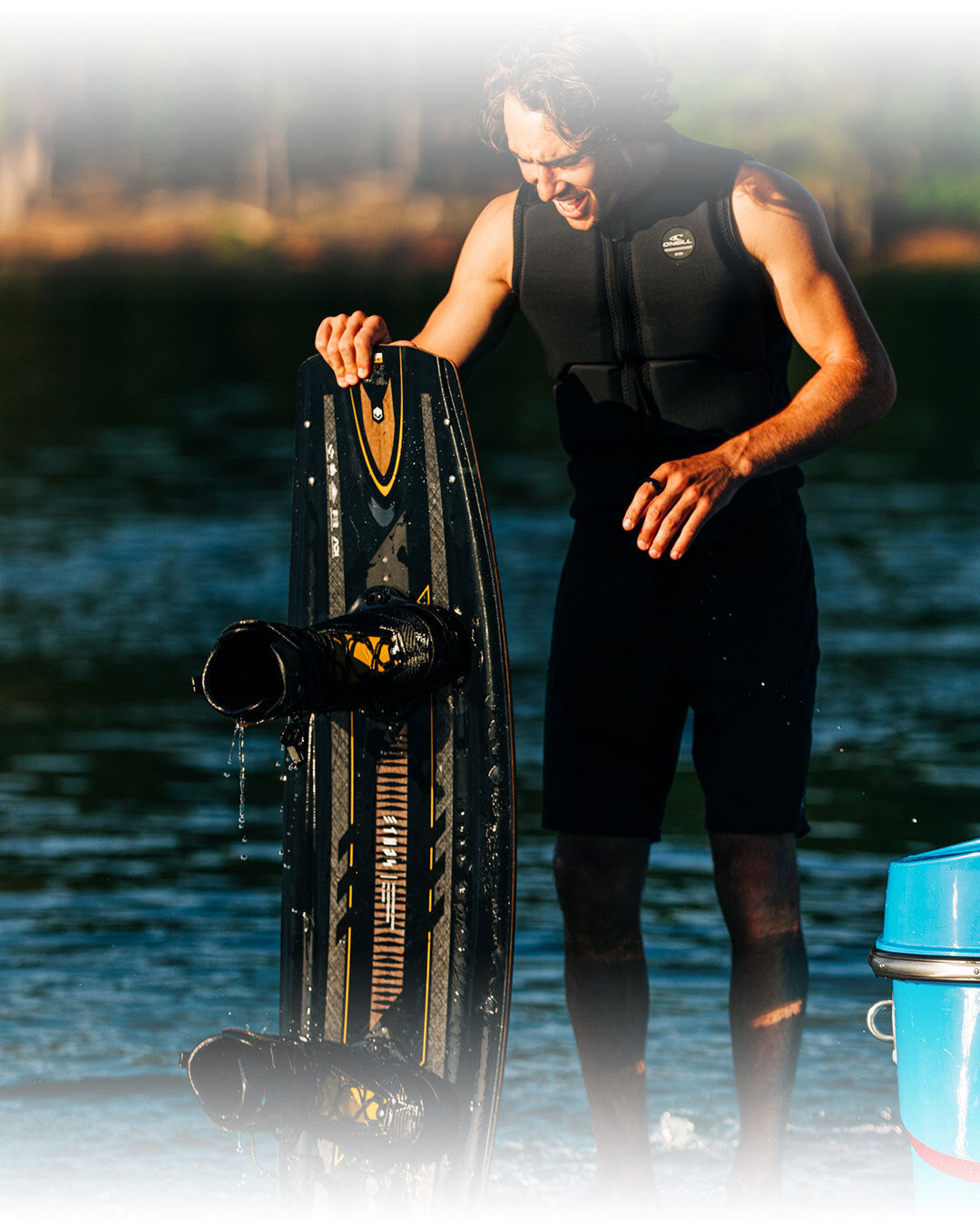 Person in a wetsuit holding a wakeboard by a body of water.