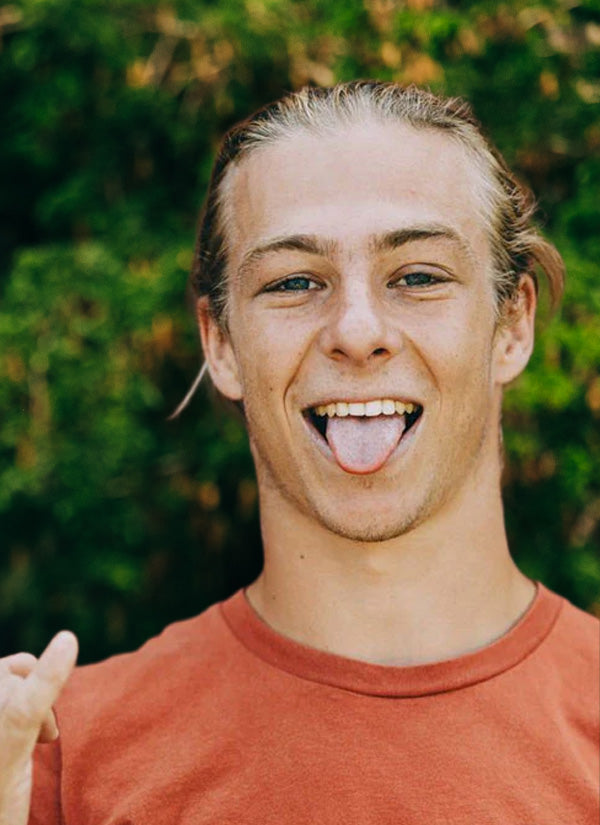 Brett P team headshot smiling with his tongue out, with long hair wearing a red shirt in front of green foliage
