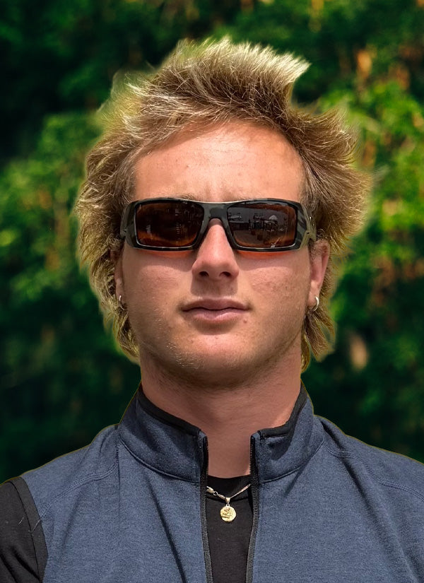 Stefano C team headshot wearing brown sunglasses, a blue vest and a necklace with trees in the background.