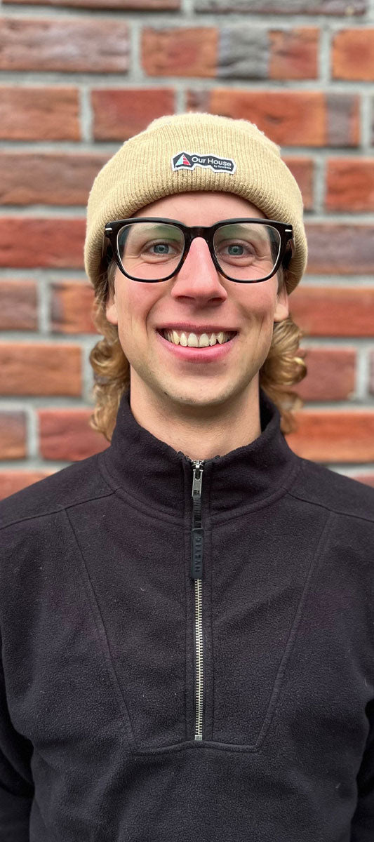 Smiling person in glasses and tan beanie stands against a red brick wall.