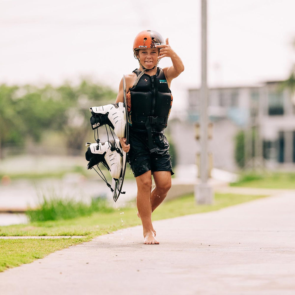 Child holding a wakeboard with white bindings and posing outdoors on a path with greenery and buildings in the background. he is wearing a black and orange helmet, a black life vest and black swimsuit.