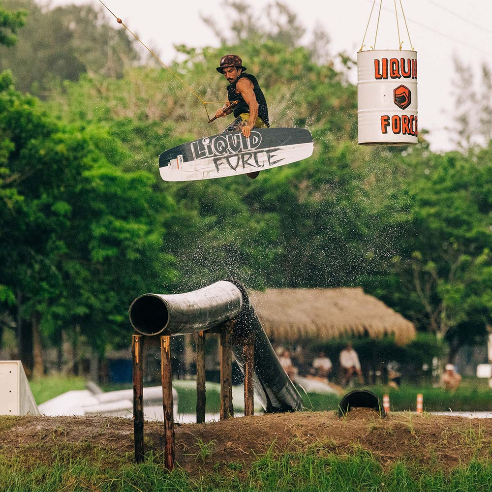 Person on a wakeboard performing a trick over a pipe with 'Liquid Force' branding on a barrel hanging from the tree, set against a natural background.