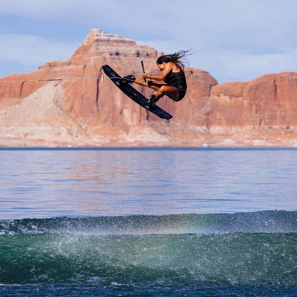 Euge D in mid-air doing a wakeboard trick over the wake with red rock cliffs in the background. She is riding a black wakeboard with a black life vest.