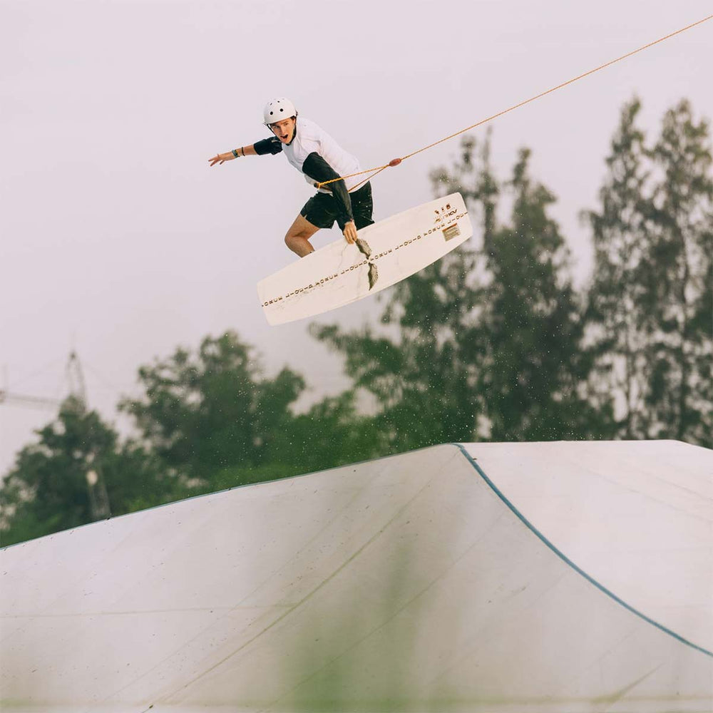Gavin S jumping over a cable park ramp riding a white wakeboard wearing a white helmet and white shirt with trees in the background