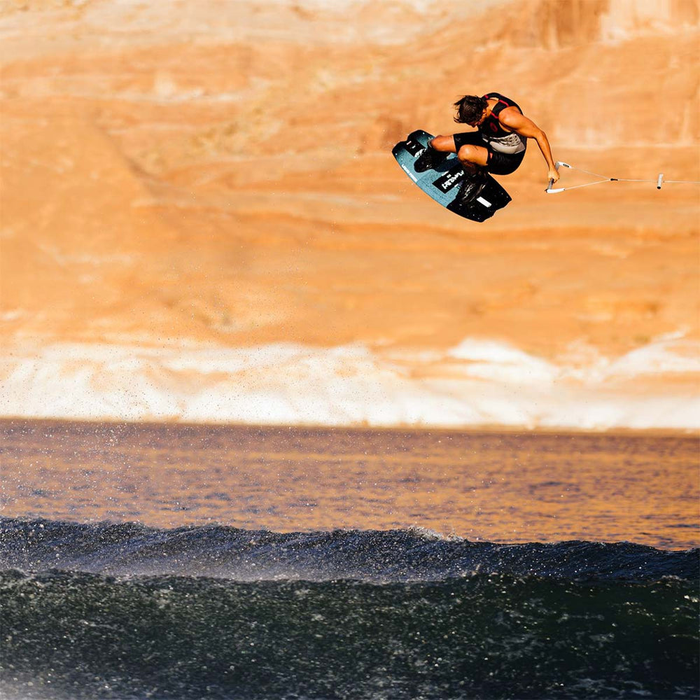 Harley C on a black and blue wakeboard performing a trick over water with a red rock background. He is wearing a black life vest and black swimsuit.