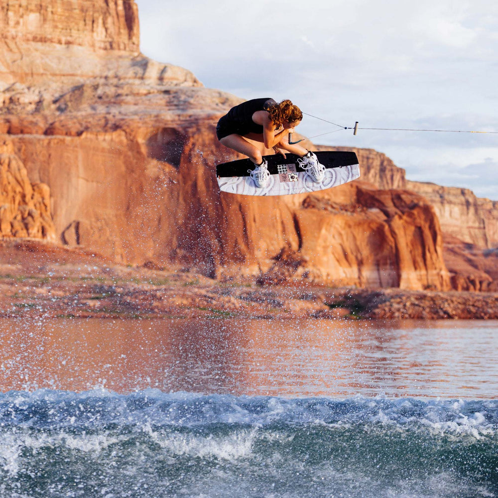 Meagan E wakeboarding jumping mid-air over water with red rock cliffs in the background. She is riding a black and white wakeboard with white bindings.