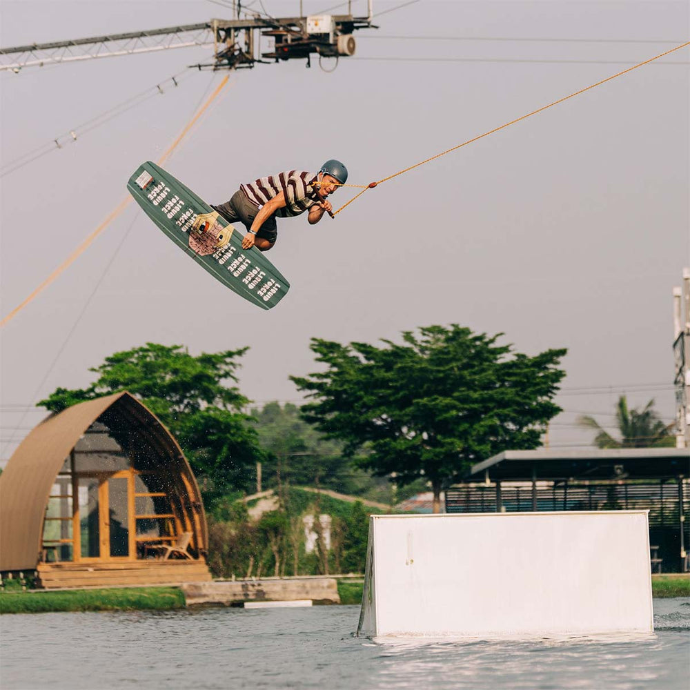 Nico V on a wakeboard performing a trick over water with trees and a building in the background. The base of the board sporting custom design and liquidForce logo. He is wearing a striped shirt and green helmet.
