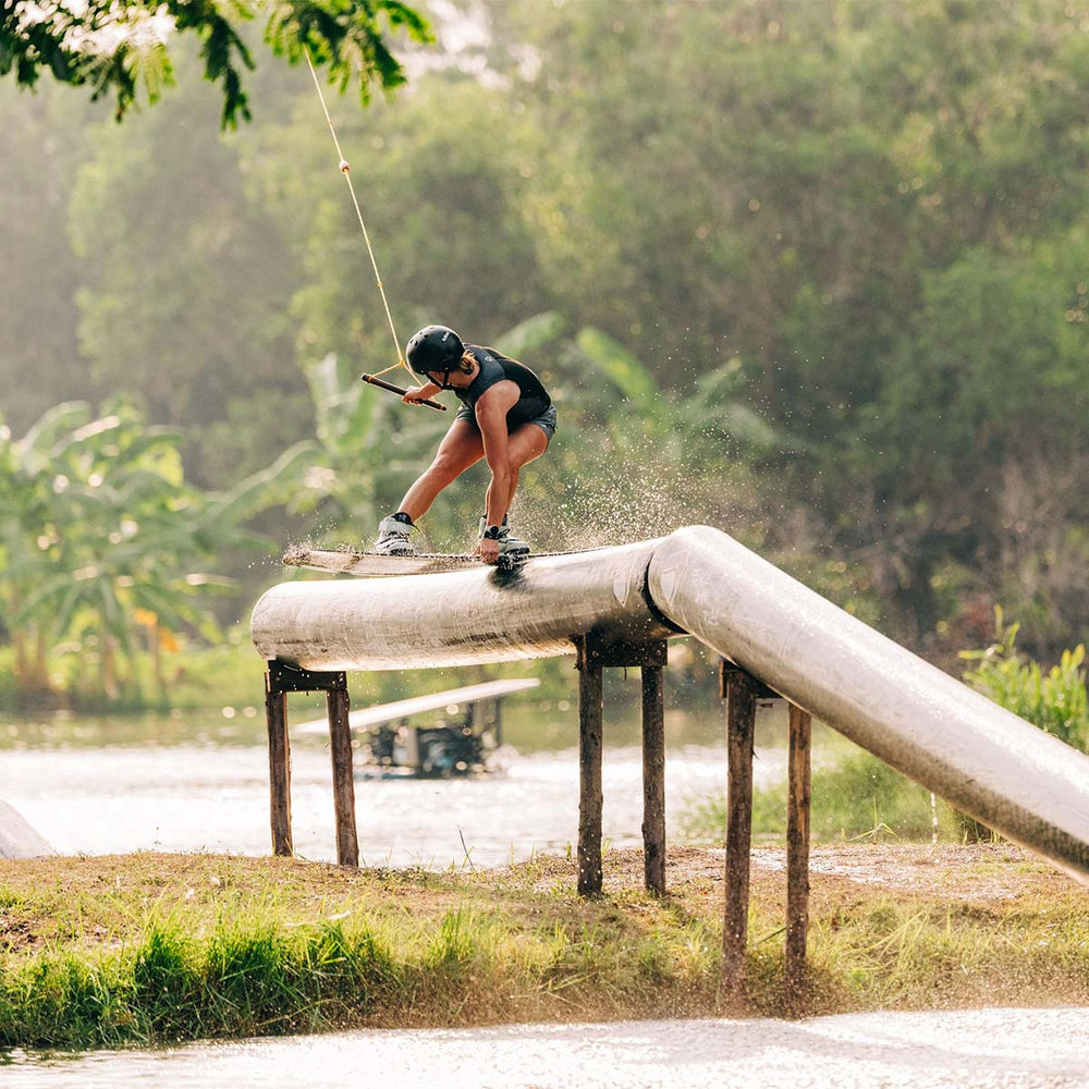 Rivers H doing a trick wakeboarding on a large pipe over dirt with a scenic background. She is wearing a black life vest and black helmet.
