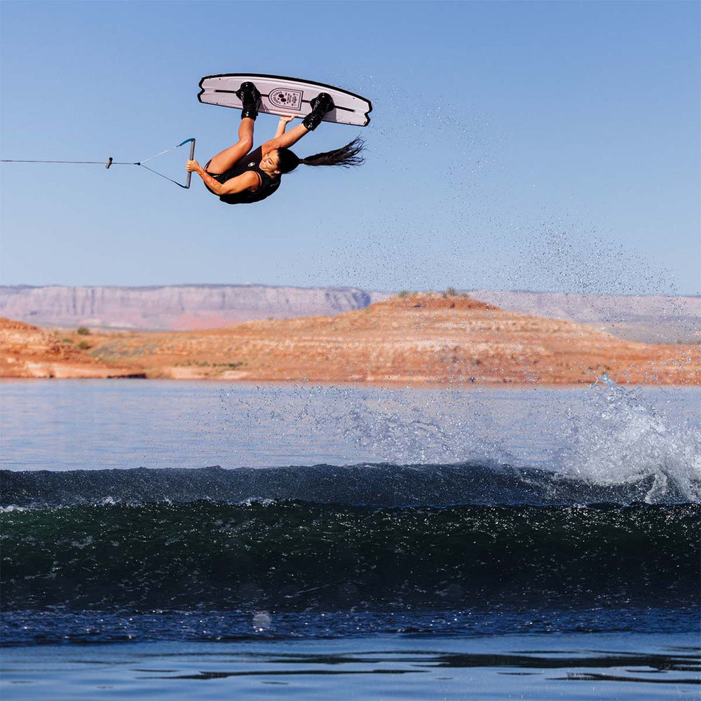Zahra K wakeboarding in mid-air flipping over a wave with a clear blue sky and distant landforms. She is riding a white and black wakeboard with black bindings.