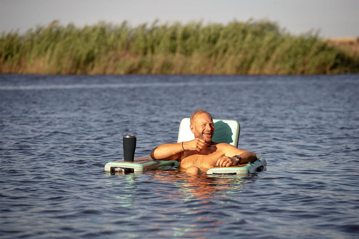 Man lounging in the water on a float chair with a drink, smiling at the camera.