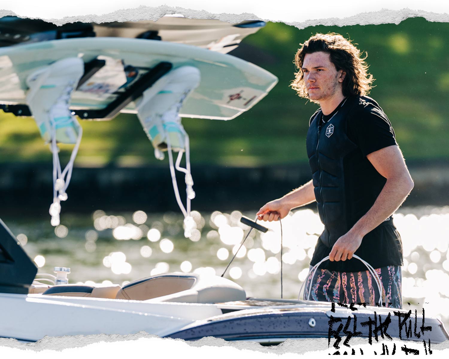 Man in black Liquid Force vest gripping a tow rope near wakeboards and a boat.