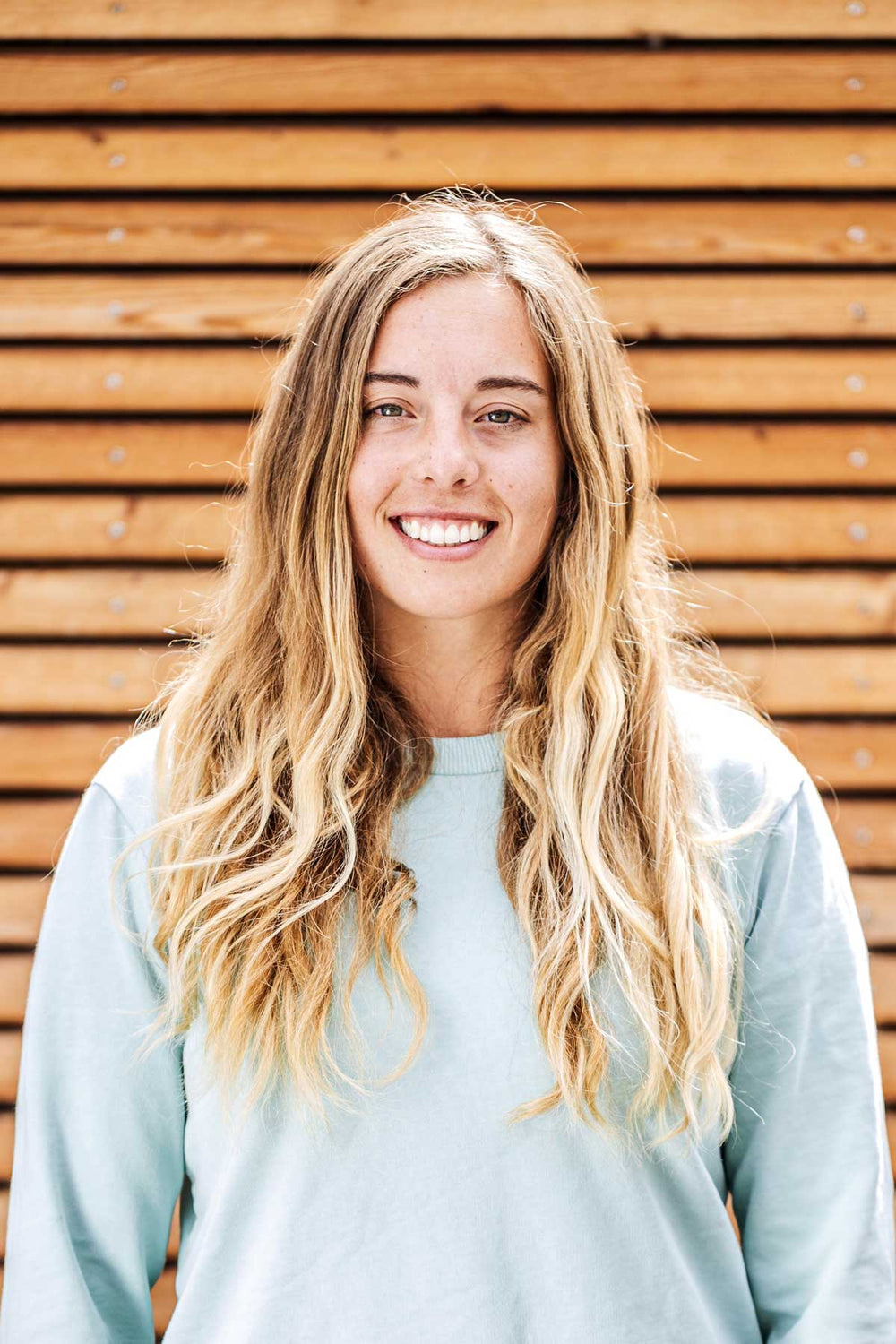 A smiling person with long hair in a light blue sweater stands against a rustic wooden wall.