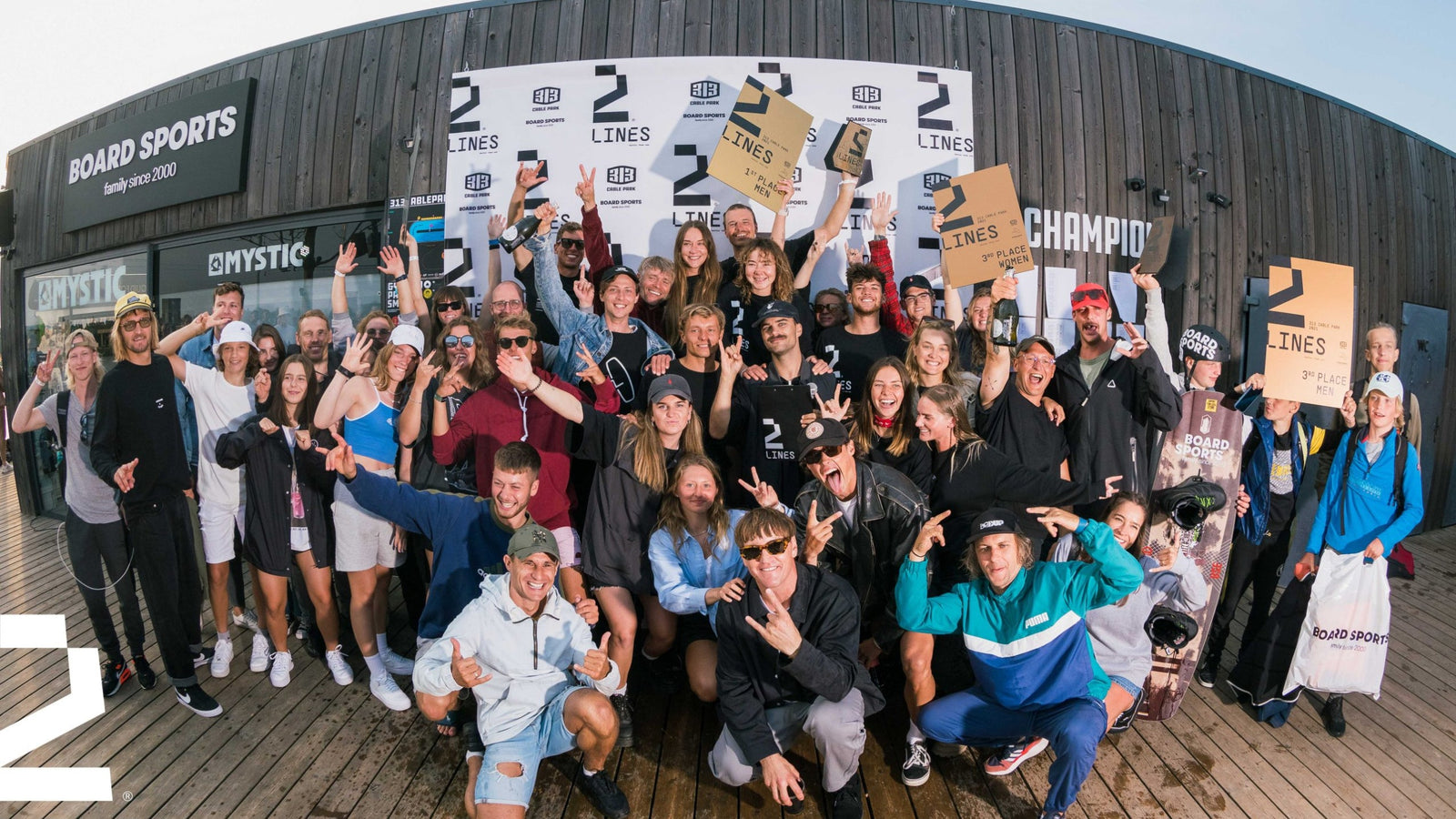 Large group celebrates on a boardwalk with trophies in front of a 2 Lines event banner.