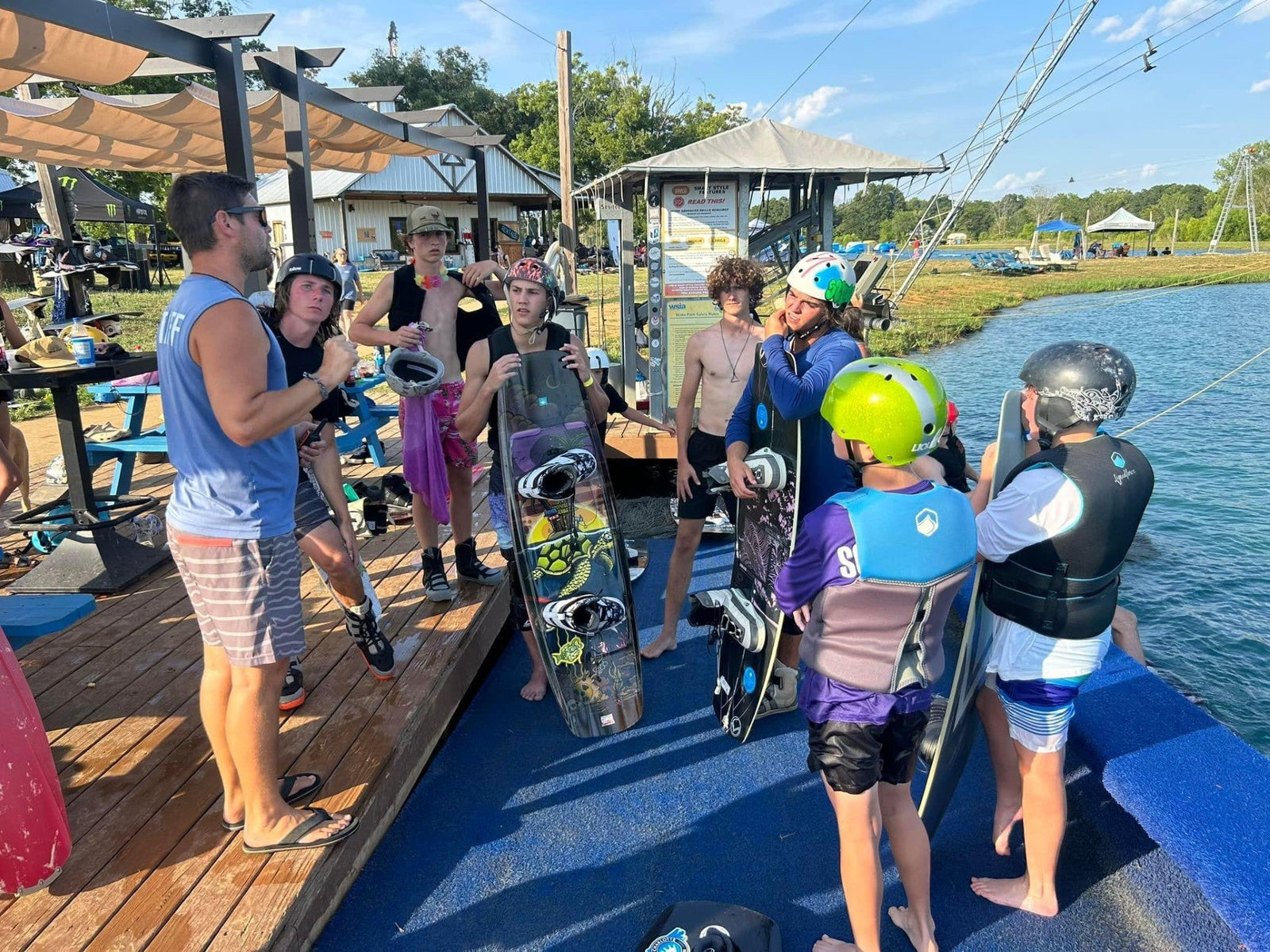 Young wakeboarders in life vests gather on dock for instruction near cable park.