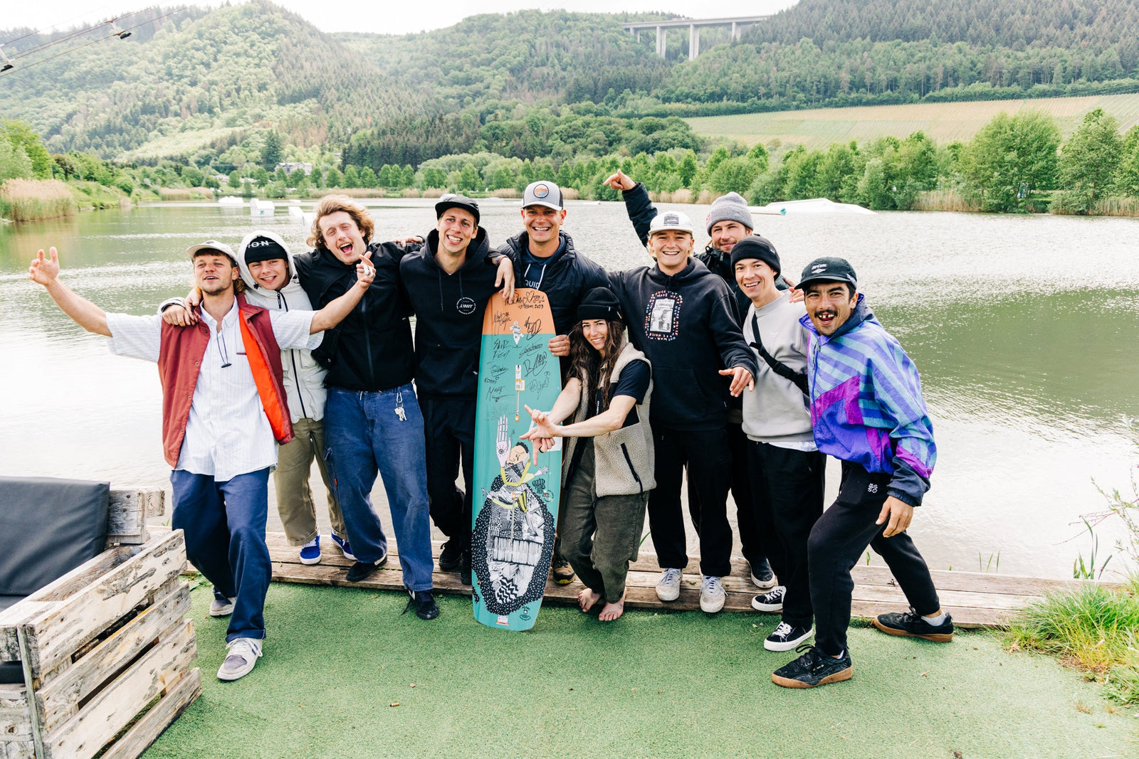 A group of friends joyfully pose by a lakeside, surrounded by lush greenery and hills.