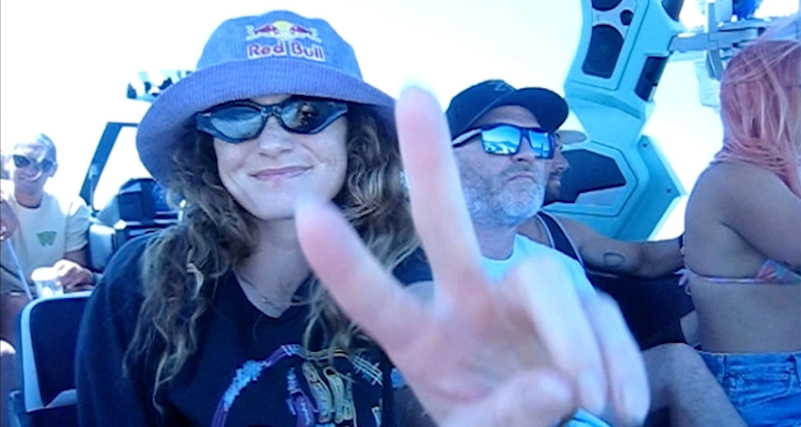 Woman in sunglasses flashes a peace sign while riding a boat with friends under bright sunlight.