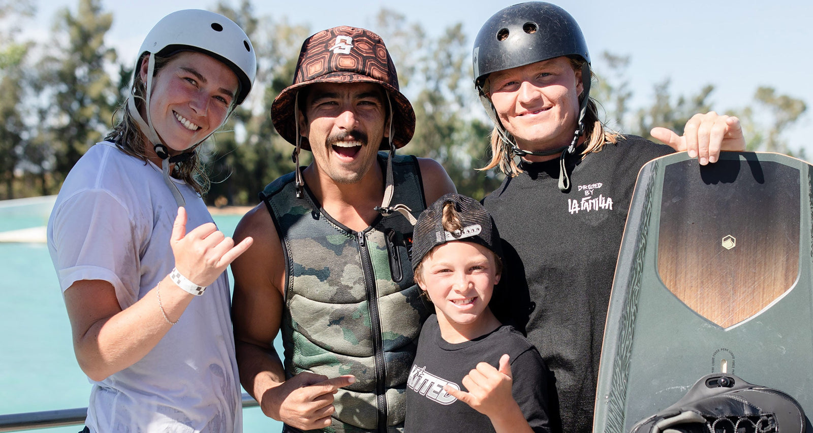 Four smiling wakeboarders pose with helmets and boards near a bright blue body of water.