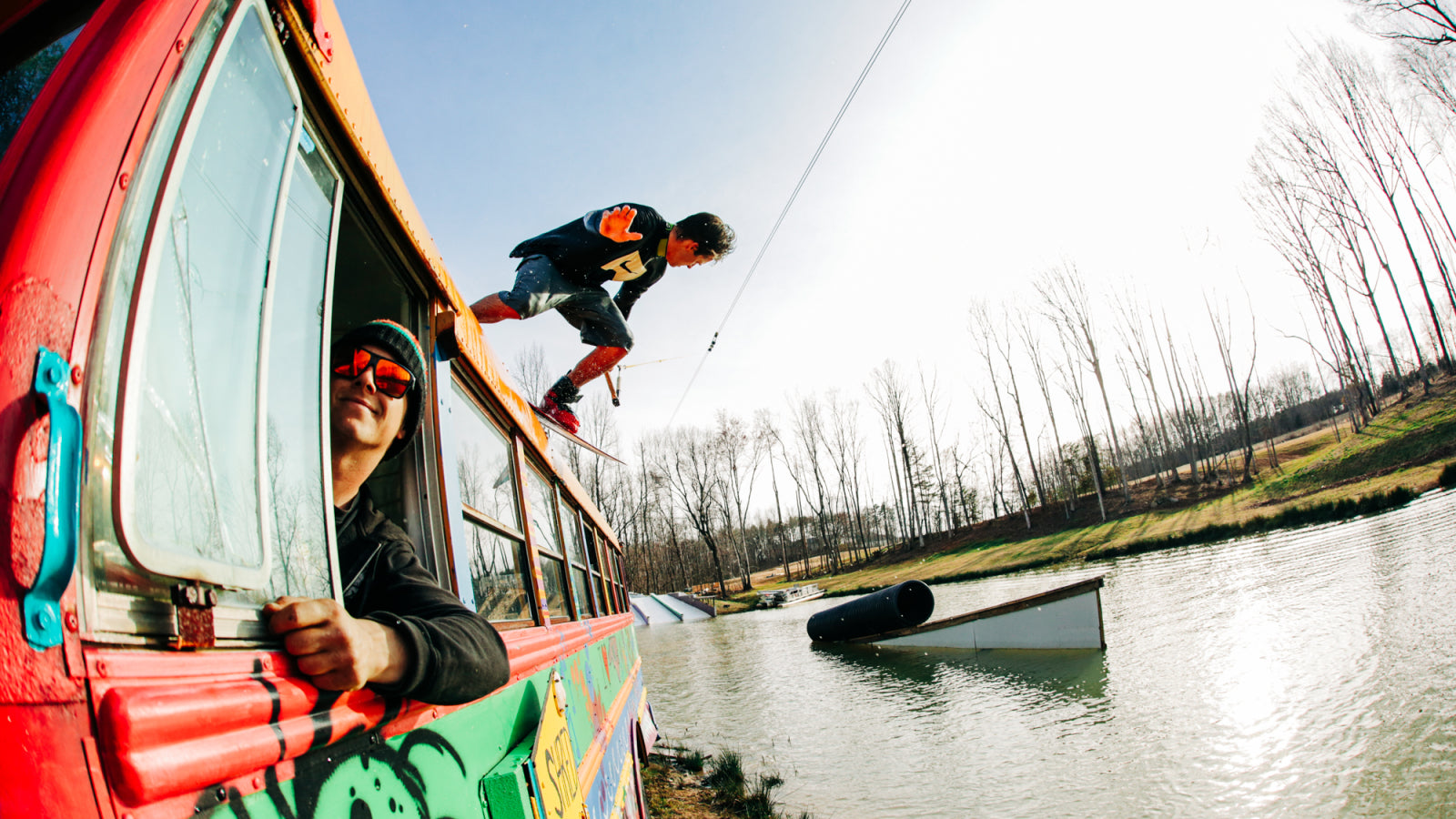 Wakeboarder jumps off colorful bus while another person leans out the window, watching.