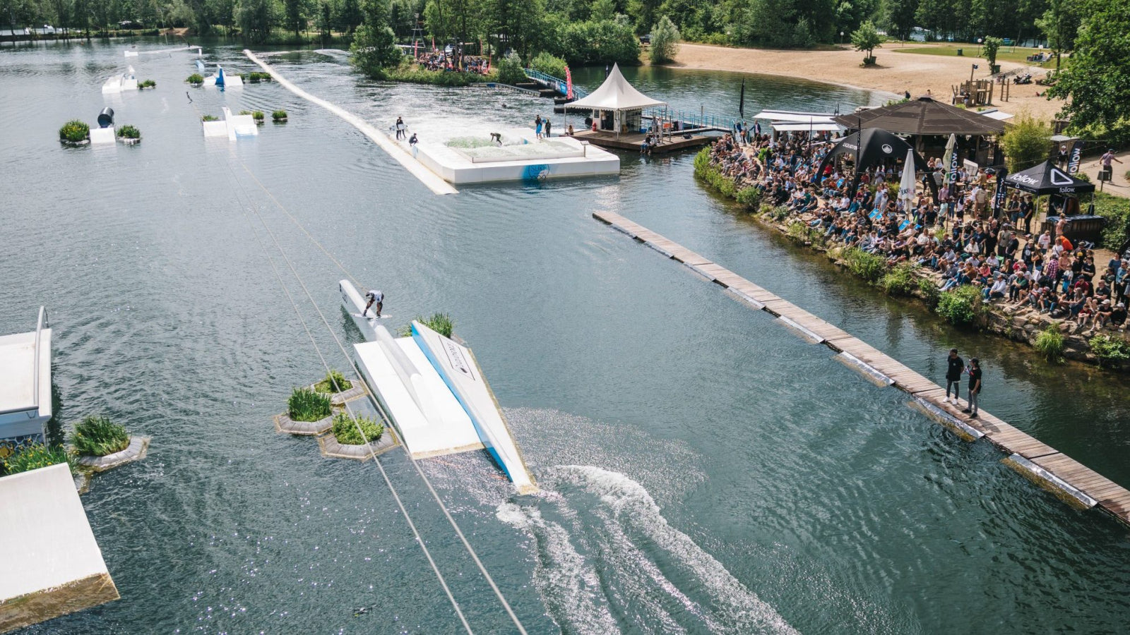 Crowd watches a wakeboarder ride a rail at a cable park event on a sunny day.