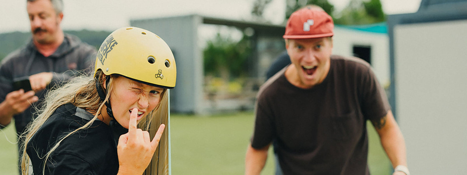 Smiling person in a yellow helmet winks and throws a hand sign next to a cheering friend.