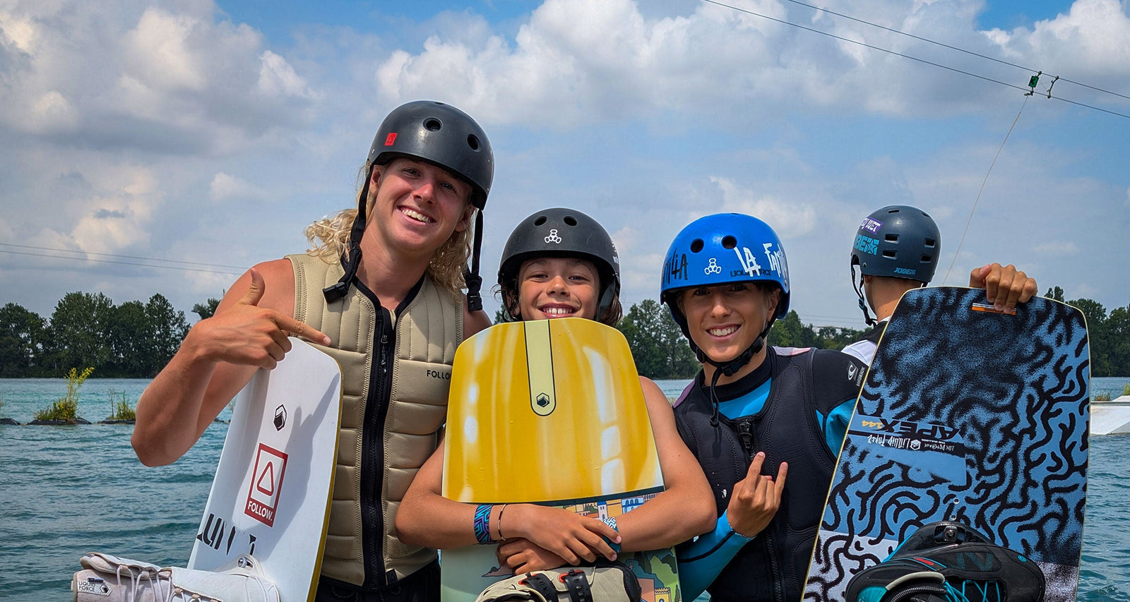 Three young wakeboarders in helmets smile on the dock holding colorful boards.