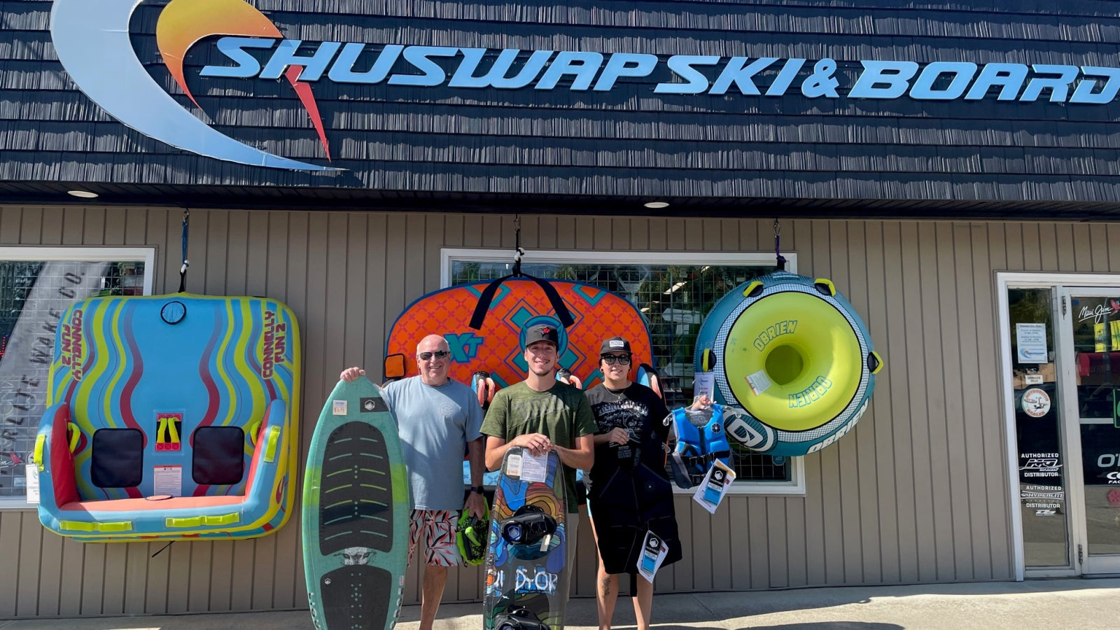 Three people holding watersports gear in front of the Shuswap Ski & Board storefront.