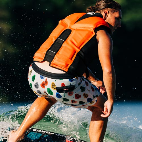 Man wakesurfing in bright orange vest and smiley face swim trunks.