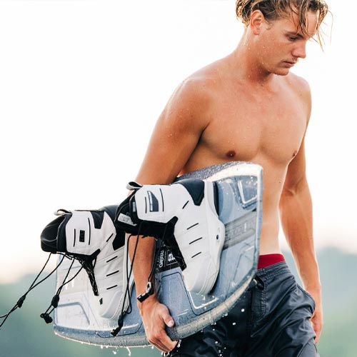 Shirtless man walking with wet gray Liquid Force wakeboard featuring white bindings.