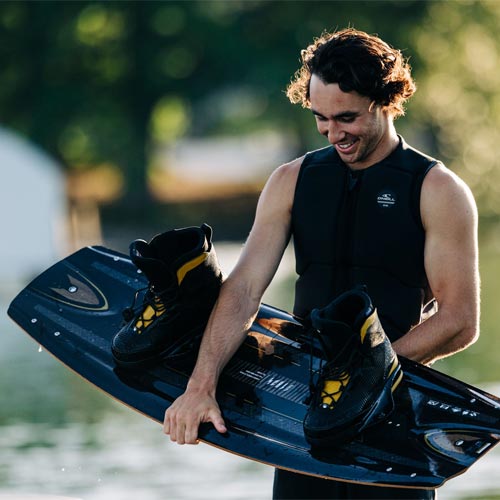 Smiling man in vest holding black Liquid Force wakeboard with yellow-accented bindings.