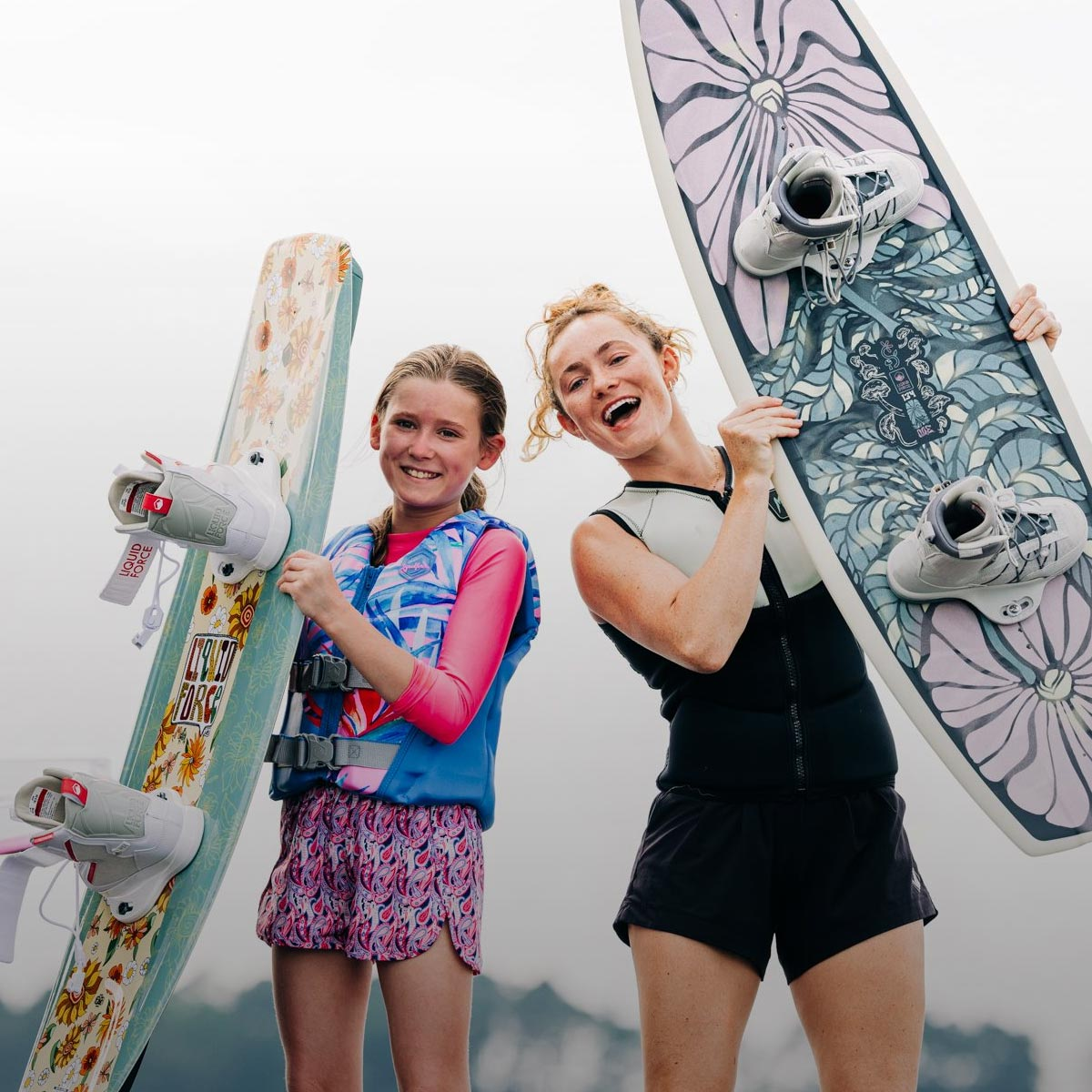 Two smiling women holding Liquid Force wakeboards with floral designs and bindings.