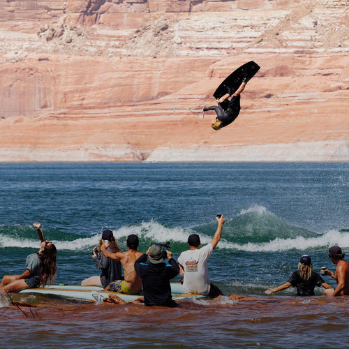 Wakeboarder mid-air performing trick above water with spectators cheering from shoreline.