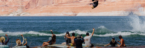 Wakeboarder mid-air performing trick above water with spectators cheering from shoreline.