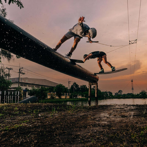 Wakeboarders grinding rail feature at sunset with cable park lines and water spray.