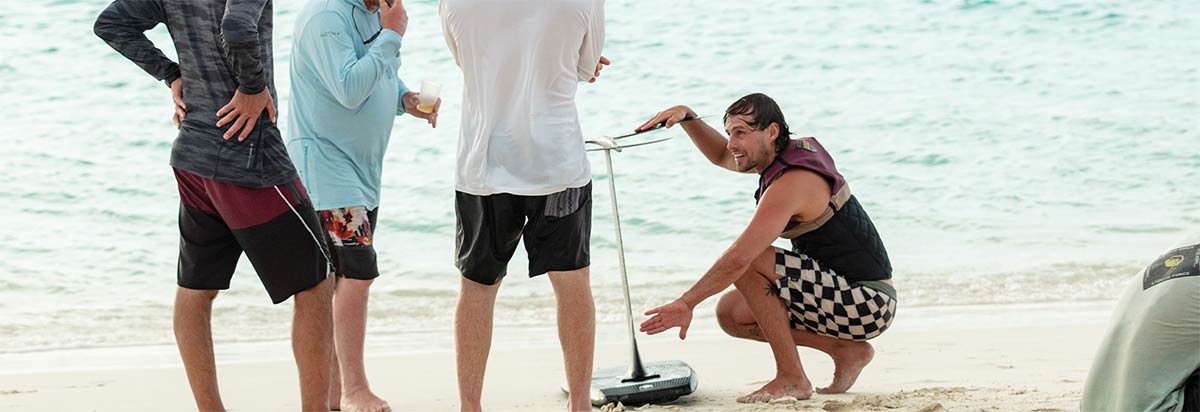 Man in a life vest crouches to explain a foil setup to three men on a sandy beach.
