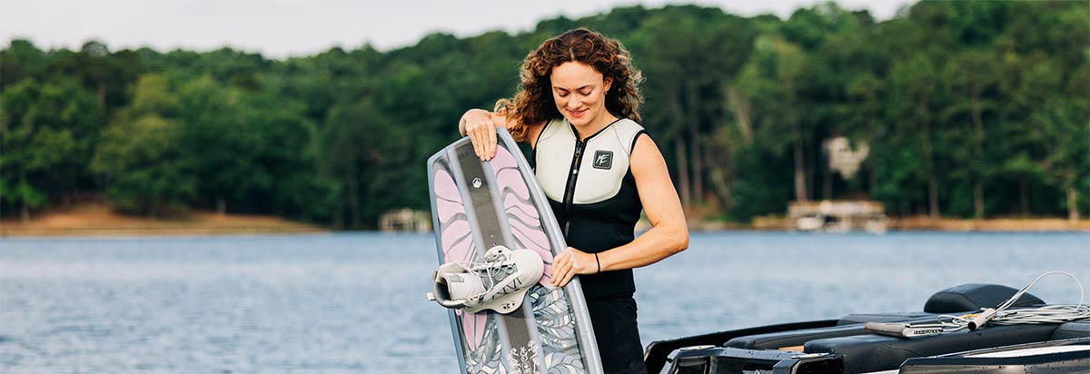 Woman stands by a boat, smiling while holding a wakeboard with mounted bindings.