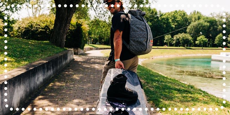 Man walking near water carrying a wakeboard with boots and wearing a backpack.