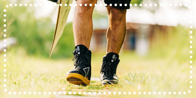 Close-up of a man walking in black wakeboard boots on a grassy path.