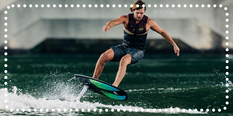 Man riding a hydrofoil board in dark water under a bridge.