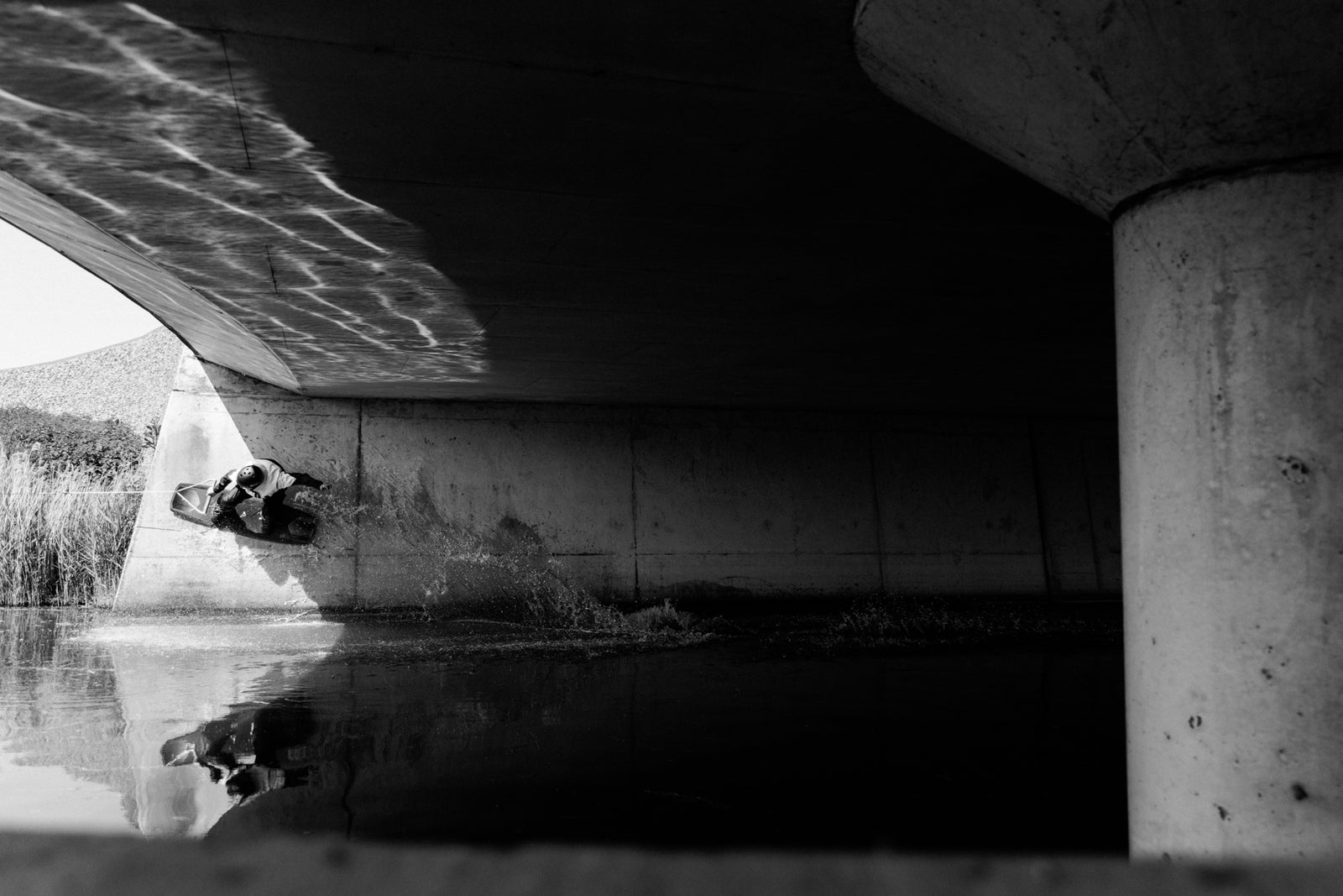 Black and white photo of a wakeboarder riding on a concrete wall under an overpass.