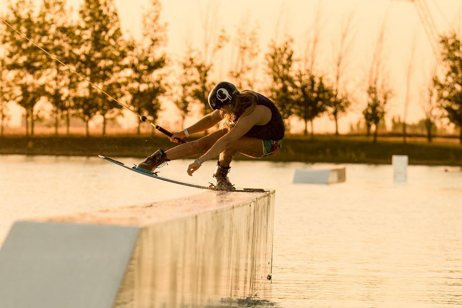 A wakeboarder balances skillfully on a narrow ramp over water during sunset.