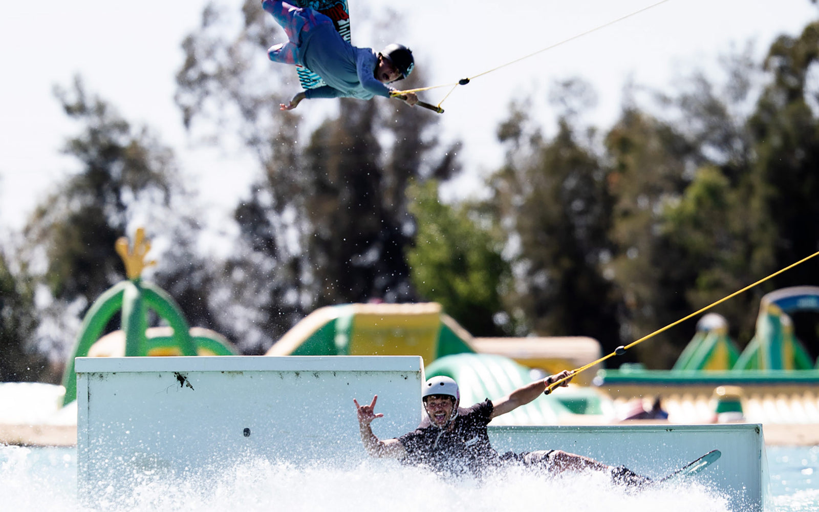 Two wakeboarders ride near a rail; one in the air, the other splashing while throwing a shaka.