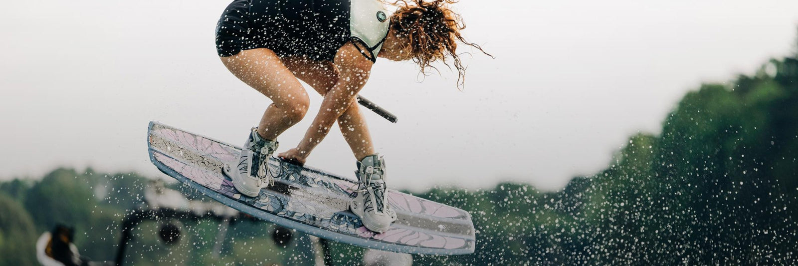 Liquid Force Boat Wakeboard Package Collection - Closeup shot of LF Athlete Meagan Ethell grabbing her board mid-air behind a wakeboard boat.