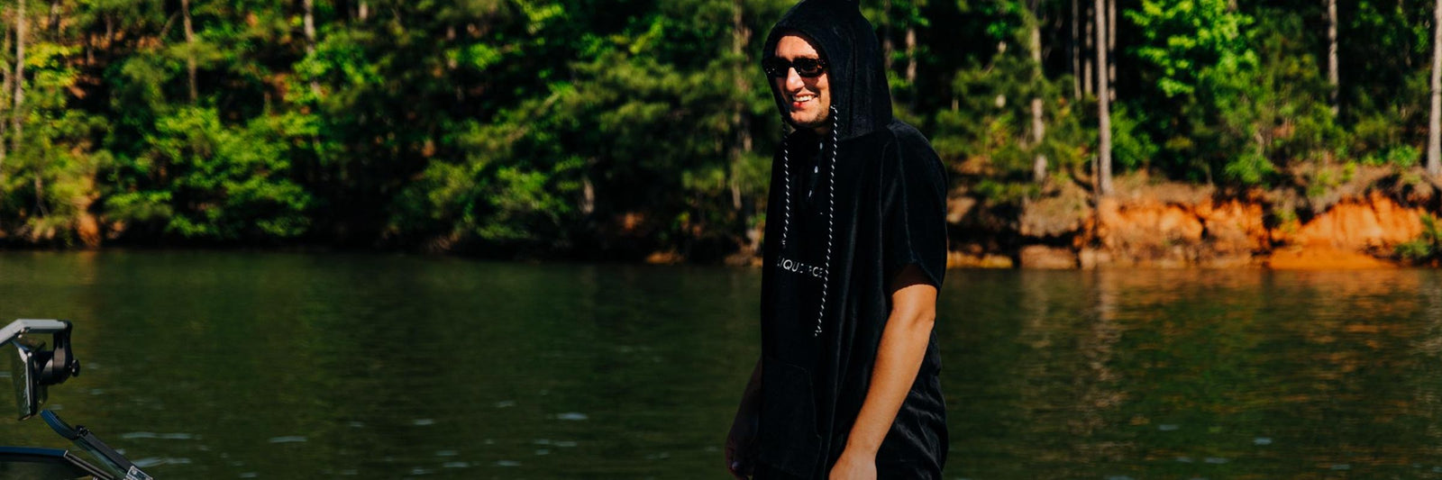 Man in a black changing poncho stands on a boat near a forested shoreline.