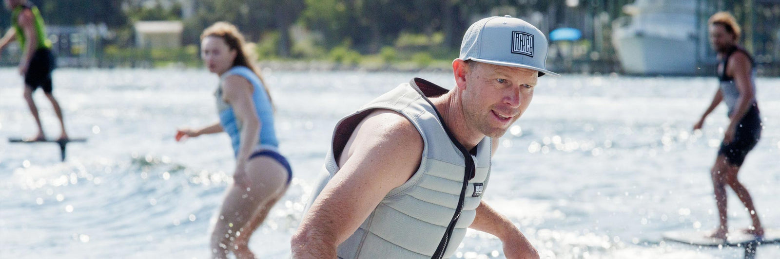 Riders foil surf on a lake, with one rider foregrounded wearing a branded hat.