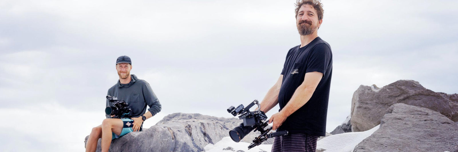 Two cameramen wearing Liquid Force shirts stand on rocky terrain with cameras.
