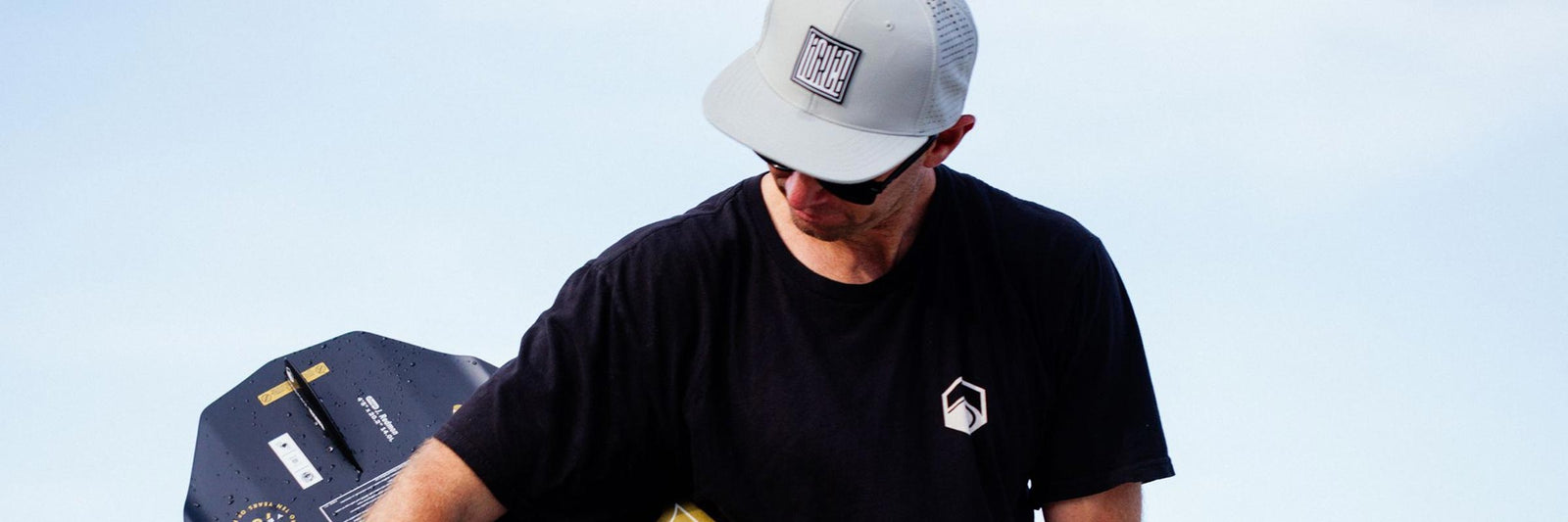 Man wearing a hat and black tee carries a wakesurf board by the water.