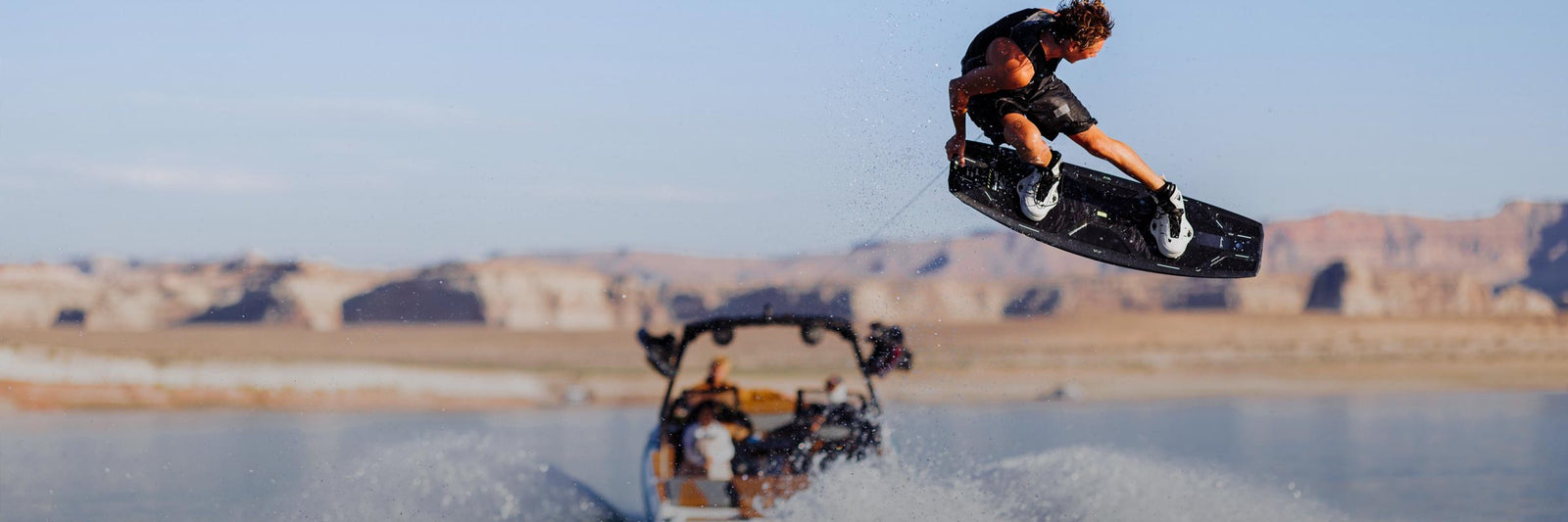 Rider launches off a wake behind a boat on a sunny lake with desert cliffs in the background.