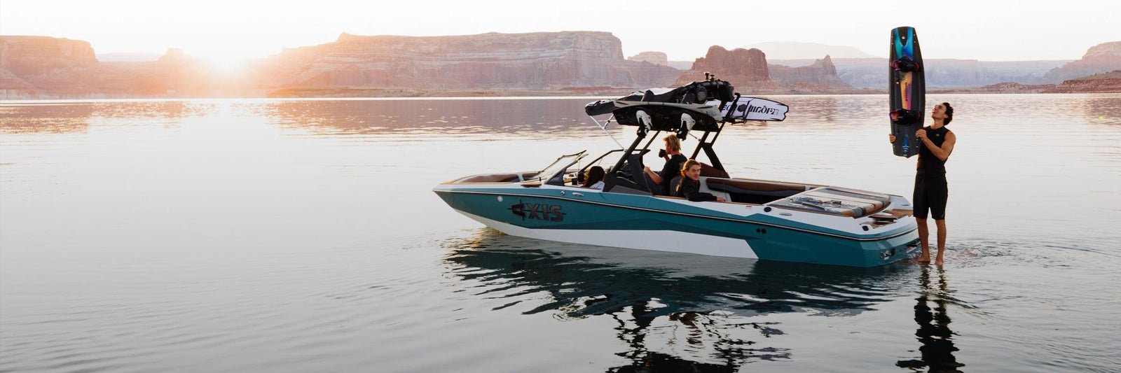 Rider holding a wakeboard beside a teal boat at sunset on a quiet lake.