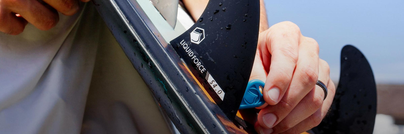 Hand installs a black Liquid Force fin onto a board using a blue tool.