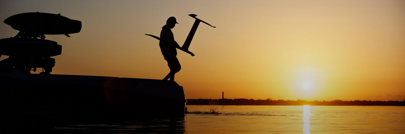Silhouette of a rider carrying a foil board beside a boat during a calm orange sunset.